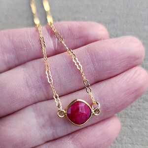 May include: A delicate gold chain necklace with a single, faceted, ruby red gemstone pendant.