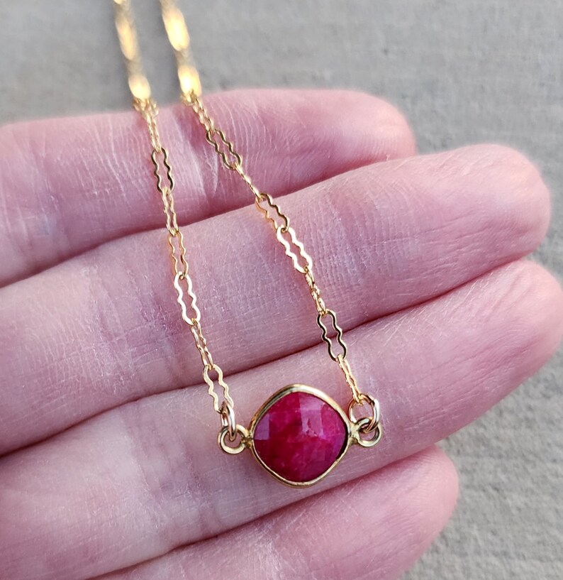 May include: A delicate gold chain necklace with a single, faceted, ruby red gemstone pendant.