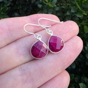 May include: A pair of drop earrings featuring faceted, teardrop-shaped deep pink gemstones set in polished silver bezels. The earrings hang from simple silver shepherd hook wires, showcasing a vibrant, translucent color and a textured surface.