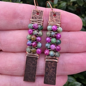 May include: Handmade earrings featuring rectangular copper-colored metal pendants and beaded strands. The beads are a mix of green, purple, and pink. The earrings are held together with copper wire and have copper-colored ear wires.