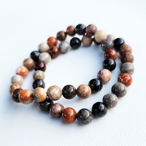 Puede incluir: Un brazalete de doble cadena hecho de cuentas de piedras preciosas marrones, negras y blancas. Las cuentas son redondas y tienen un acabado natural y pulido.