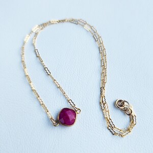 May include: A delicate gold chain necklace with a single, faceted, deep red gemstone pendant.