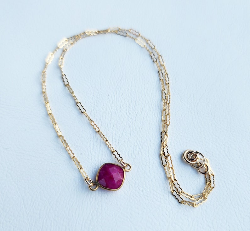 May include: A delicate gold chain necklace with a single, faceted, deep red gemstone pendant.