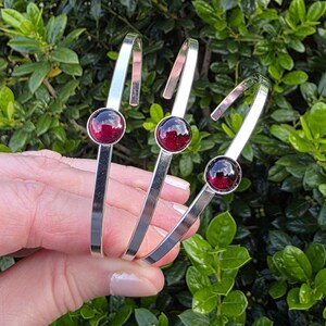 May include: Three silver-toned bracelets with a polished finish, each featuring a round, deep red gemstone. The bracelets are held against a backdrop of green foliage, showcasing their design and color.