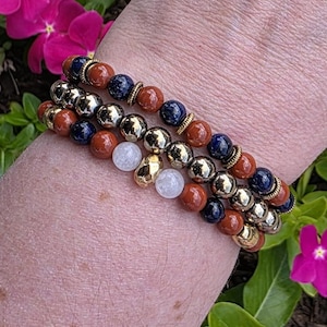 May include: A stack of three beaded bracelets featuring alternating round beads in dark blue, reddish-brown, and metallic gold tones. The design includes small gold-colored spacers and two translucent white beads positioned in the center of the stack.