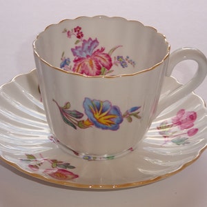 May include: A white porcelain teacup and saucer with a floral design. The cup has a scalloped rim and a gold trim. The saucer has a scalloped edge and a pink flower design.