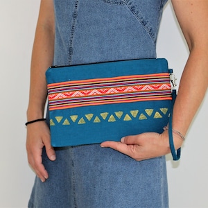 May include: Teal clutch with a colorful woven band across the top and gold triangle accents. The clutch has a black zipper and a wrist strap. The woven band features red, orange, and white geometric patterns.