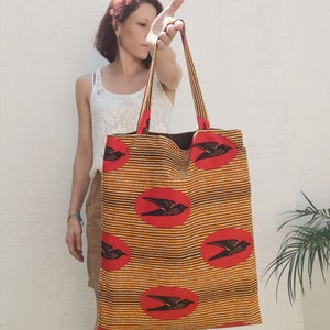May include: A large tote bag with a repeating pattern of black birds in red circles on a yellow and black striped background. The bag has long straps and a brown interior.