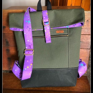 May include: A green and black canvas backpack with a purple and multi-colored patterned strap. The backpack has a flap with a zippered pocket and a small leather patch that says "Tiny Wren Goods".
