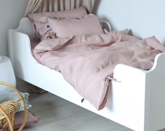Könnte beinhalten: Ein weißes Kinderbett aus Holz mit einer rosafarbenen Bettwäsche aus Leinen und zwei Kissen. Das Bett hat ein abgerundetes Kopfteil und Fußteil.
