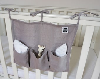 Könnte beinhalten: Ein grauer Bettorganizer aus Leinen mit vier Taschen, der an einem weißen Babybett hängt. Der Organizer hat ein weißes Etikett mit dem Text "NURKO". In den Taschen befinden sich weiße Tücher und ein beiges Giraffenspielzeug.