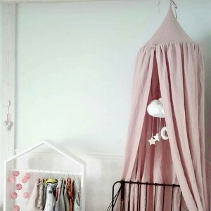 Puede incluir: Dormitorio infantil con una cama con dosel rosa, un perchero blanco en forma de casa y una colcha con un colorido estampado de &aacute;rboles. Un m&oacute;vil con nubes y estrellas cuelga del dosel.