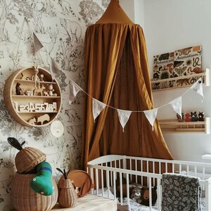 May include: A nursery interior with a white cot, wooden chest of drawers, and a round shelf displaying toys and numbers. A mustard-coloured canopy hangs above the cot. The room features woodland animal wallpaper and decorative elements.