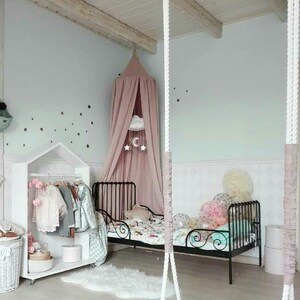 Puede incluir: Un dormitorio infantil con una combinaci&oacute;n de colores blanco y rosa. La habitaci&oacute;n cuenta con una cama de metal negro, un armario blanco con ropa, un columpio y un dosel sobre la cama. Estrellas decoran la pared.