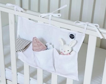 Könnte beinhalten: Ein weißer Stoff-Bettorganizer mit drei Taschen, der mit weißen Bändern am Bett befestigt wird. Die Taschen enthalten einen gestreiften Stoffartikel, einen rosa blattförmigen Artikel und ein weißes Hasen-Plüschtier. Der Organizer hat ein schwarz-weißes "MUKKO"-Etikett.