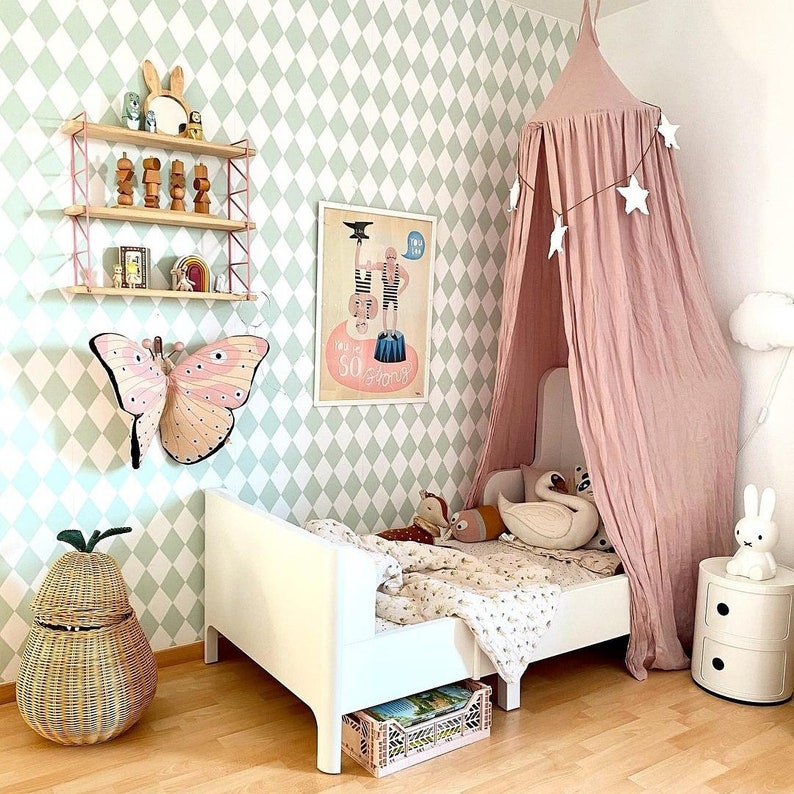 Puede incluir: Dormitorio infantil con una cama blanca, un dosel rosa y una pared decorada con una gran mariposa y un p&oacute;ster. La habitaci&oacute;n tiene un papel pintado con estampado de rombos verde claro y blanco, una cesta en forma de pera de mimbre y una mesita de noche blanca.