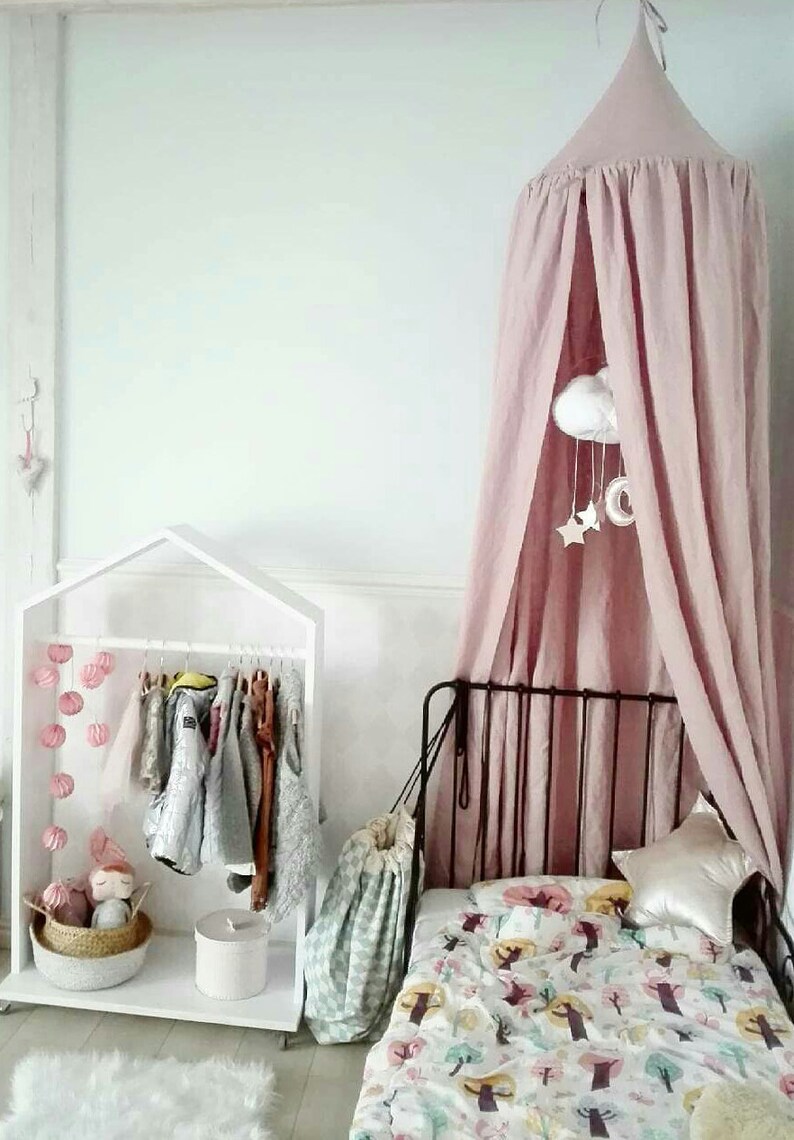Puede incluir: Dormitorio infantil con una cama con dosel rosa, un perchero blanco en forma de casa y una colcha con un colorido estampado de &aacute;rboles. Un m&oacute;vil con nubes y estrellas cuelga del dosel.