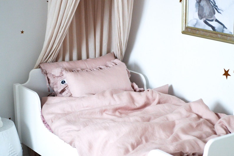 May include: A white bed frame with a pink linen duvet and two pink pillows. The bed has a canopy made of sheer fabric.