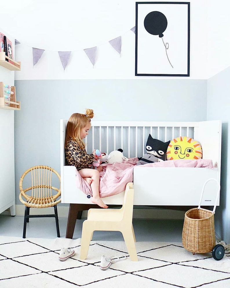May include: A white crib with a pink blanket and a few stuffed animals. A child is sitting on a wooden chair next to the crib. There is a wicker basket and a black and white rug on the floor.