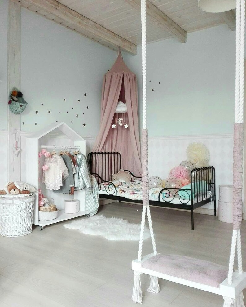 Puede incluir: Un dormitorio infantil con una combinaci&oacute;n de colores blanco y rosa. La habitaci&oacute;n cuenta con una cama de metal negro, un armario blanco con ropa, un columpio y un dosel sobre la cama. Estrellas decoran la pared.