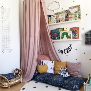 Puede incluir: Un rinc&oacute;n de lectura infantil con un dosel rosa, cojines y un colch&oacute;n de suelo. Una peque&ntilde;a cama de rat&aacute;n con un mono de peluche est&aacute; al lado. Estanter&iacute;as y una l&aacute;mpara con forma de nube est&aacute;n en la pared, con una alfombra blanca con lunares negros.