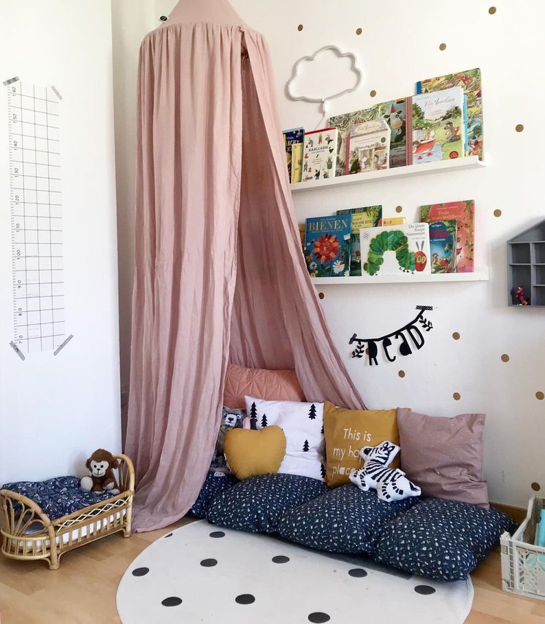 Puede incluir: Un rinc&oacute;n de lectura infantil con un dosel rosa, cojines y un colch&oacute;n de suelo. Una peque&ntilde;a cama de rat&aacute;n con un mono de peluche est&aacute; al lado. Estanter&iacute;as y una l&aacute;mpara con forma de nube est&aacute;n en la pared, con una alfombra blanca con lunares negros.