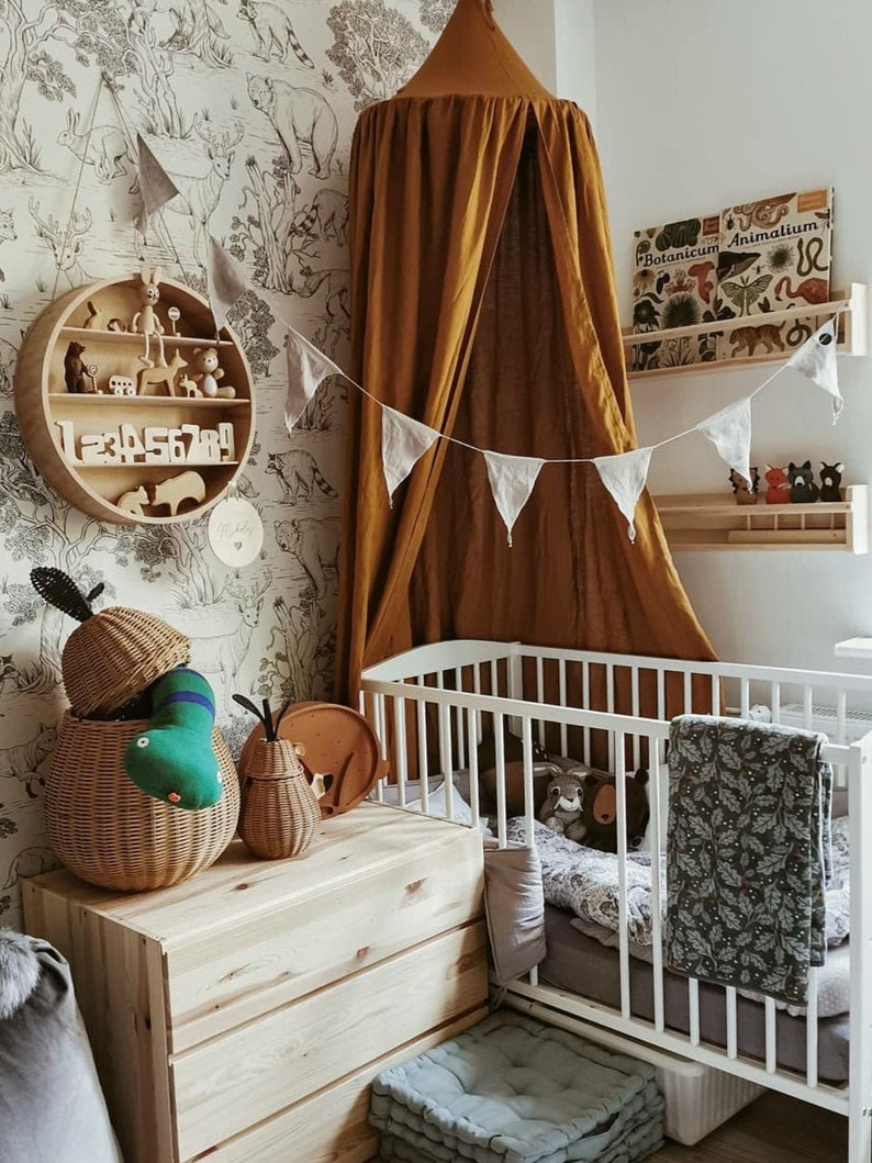 May include: A nursery interior with a white cot, wooden chest of drawers, and a round shelf displaying toys and numbers. A mustard-coloured canopy hangs above the cot. The room features woodland animal wallpaper and decorative elements.