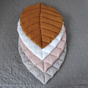 May include: Four quilted leaf-shaped play mats in different colors: brown, white, pink, and gray. The mats are stacked on top of each other.