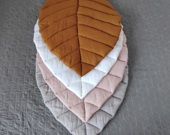 Puede incluir: Cuatro colchonetas de juego acolchadas en forma de hoja en diferentes colores: marrón, blanco, rosa y gris. Las colchonetas están apiladas unas sobre otras.