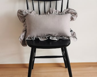 Könnte beinhalten: Ein graues Leinenkissen mit Rüschen sitzt auf einem schwarzen Holzstuhl. Der Stuhl hat eine geschwungene Rückenlehne und ein schlichtes Design.