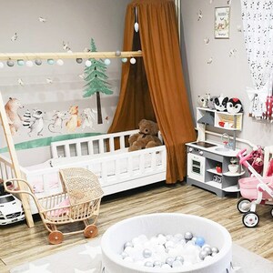 May include: A children's playroom with a white cot, a brown canopy, and a ball pit. The room features a wooden play kitchen, a toy pram, and a wall mural with forest animals. The floor is light wood, and the walls are grey.