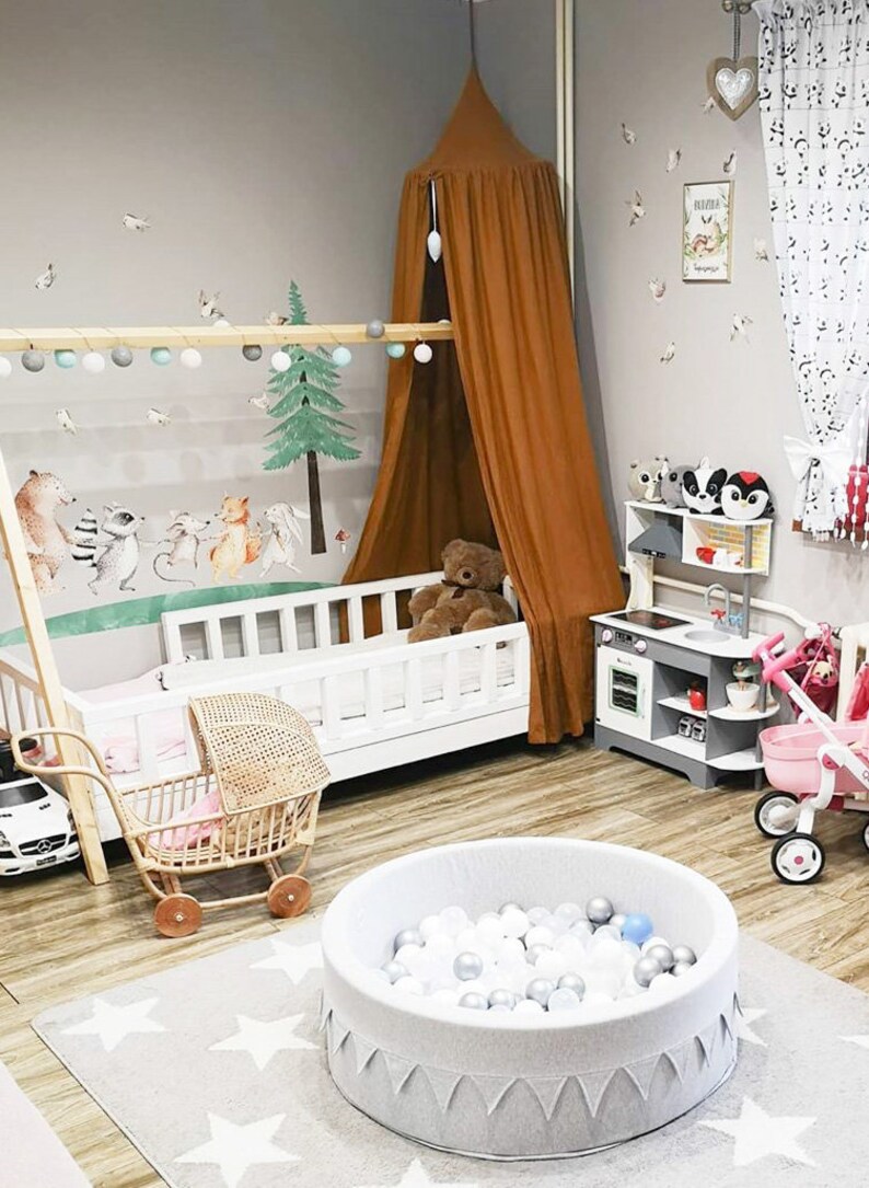 May include: A children's playroom with a white cot, a brown canopy, and a ball pit. The room features a wooden play kitchen, a toy pram, and a wall mural with forest animals. The floor is light wood, and the walls are grey.