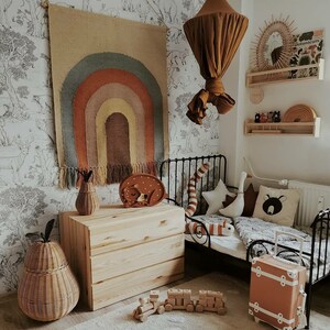 May include: A child's bedroom with a neutral colour palette. A woven rainbow wall hanging, a wooden chest of drawers, and a small bed with decorative pillows. A small orange suitcase and a wooden toy train are on the floor.