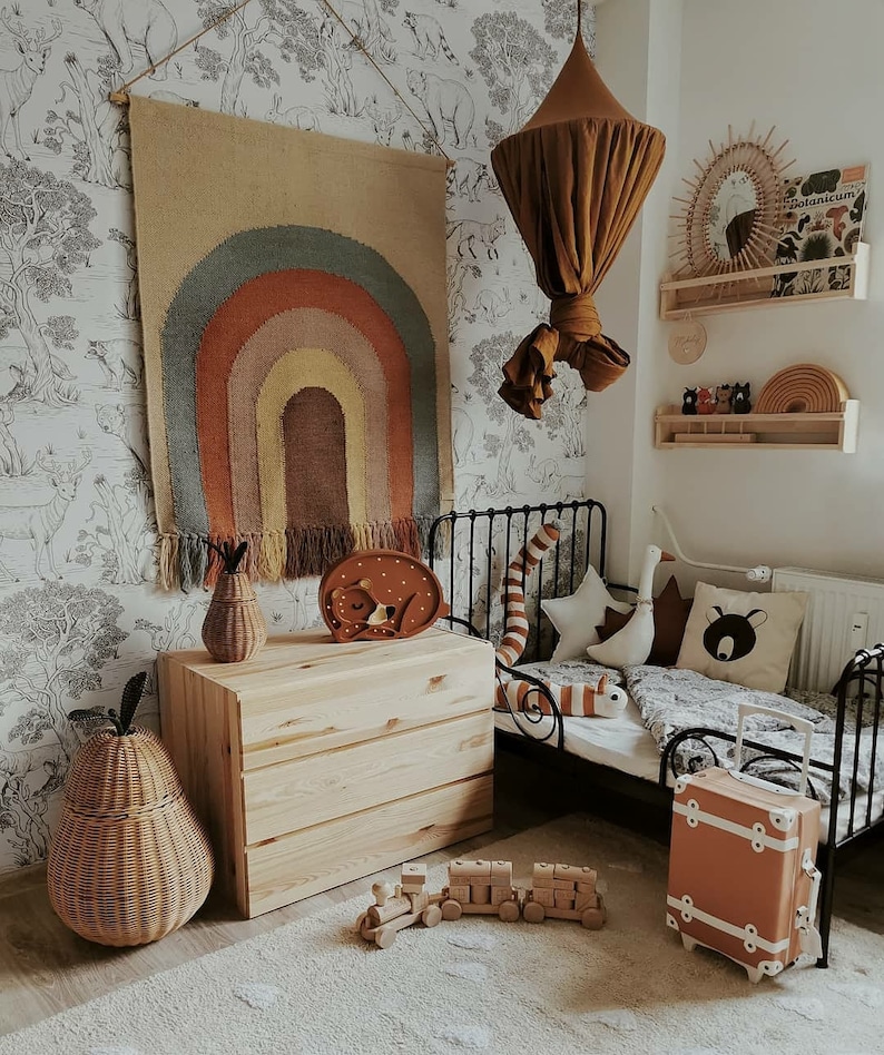 May include: A child's bedroom with a neutral colour palette. A woven rainbow wall hanging, a wooden chest of drawers, and a small bed with decorative pillows. A small orange suitcase and a wooden toy train are on the floor.