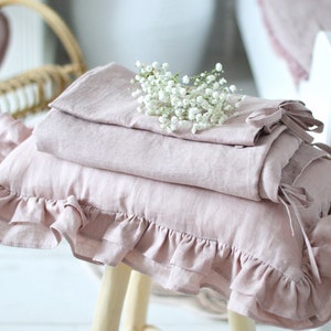 May include: A stack of pink linen pillowcases with ruffled edges, one with a bow, on a wooden stool with a bouquet of white baby's breath flowers on top.