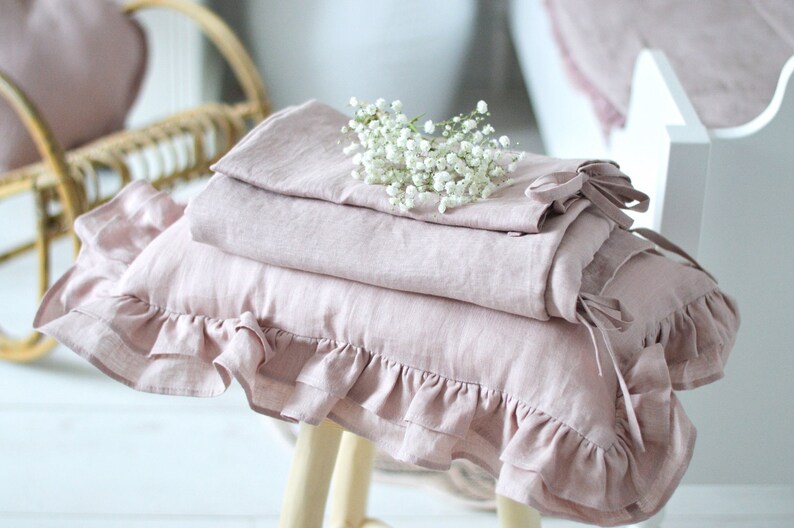 May include: A stack of pink linen pillowcases with ruffled edges, one with a bow, on a wooden stool with a bouquet of white baby's breath flowers on top.