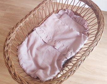 Könnte beinhalten: Ein gewebter Weidenkorb-Stubenwagen mit einer weichen, hellvioletten Babydecke. Die Decke hat einen gerüschten Rand und liegt in dem oval geformten Korb. Der Stubenwagen ist aus naturfarbenem Weidengeflecht.