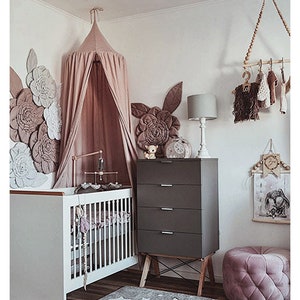 Puede incluir: Un cuarto de beb&eacute; con una cuna blanca, una c&oacute;moda gris y un dosel rosa. La pared presenta grandes decoraciones florales en tonos rosas y blancos. Una peque&ntilde;a alfombra y un puf rosa completan la decoraci&oacute;n de la habitaci&oacute;n.