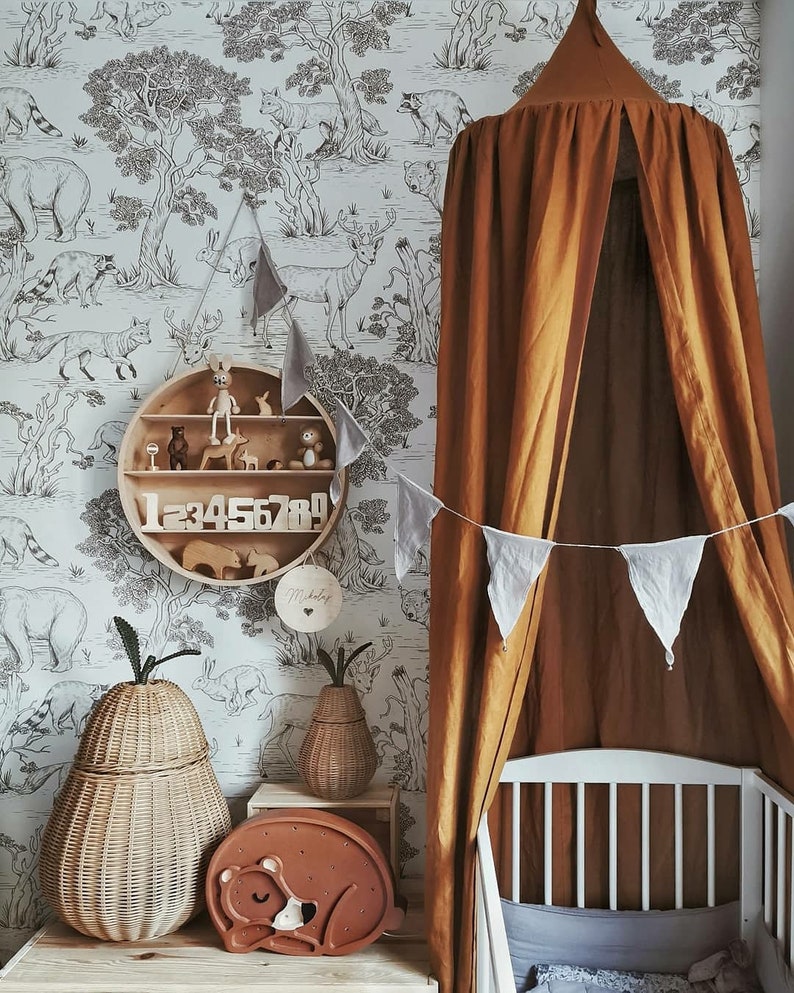 May include: A nursery interior featuring a white cot, a brown canopy, and a wooden shelf with toys and numbers. The room has a wallpaper with a black and white forest design. Wicker pear-shaped baskets and a bear-shaped night light complete the decor.