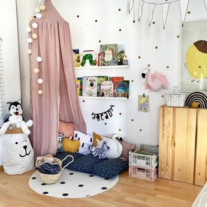 Puede incluir: Un dormitorio infantil con un dosel rosa, estanter&iacute;as blancas con libros y una pared blanca decorada con lunares dorados. Un perro husky de peluche est&aacute; sentado encima de una bolsa de almacenamiento blanca con forma de oso. Una alfombra redonda con lunares negros est&aacute; en el suelo.