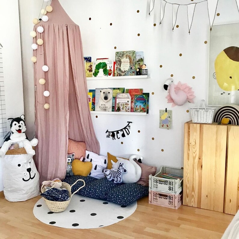 Puede incluir: Un dormitorio infantil con un dosel rosa, estanter&iacute;as blancas con libros y una pared blanca decorada con lunares dorados. Un perro husky de peluche est&aacute; sentado encima de una bolsa de almacenamiento blanca con forma de oso. Una alfombra redonda con lunares negros est&aacute; en el suelo.