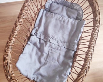 Könnte beinhalten: Ein Baby-Stubenwagen aus geflochtenem Weidenholz mit einem grauen Leinen-Bettwäsche-Set. Die Bettwäsche umfasst ein Kissen und eine Decke mit Rüschenkanten. Der ovale Stubenwagen steht auf einem Holzboden.