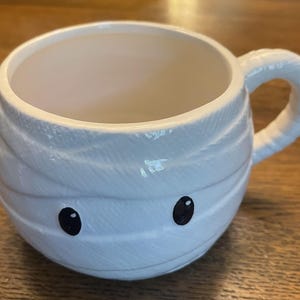 Puede incluir: Taza de cer&aacute;mica blanca con forma de momia con ojos negros. La taza tiene un asa y es perfecta para Halloween.