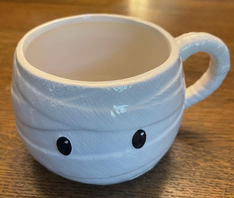 Puede incluir: Taza de cer&aacute;mica blanca con forma de momia con ojos negros. La taza tiene un asa y es perfecta para Halloween.