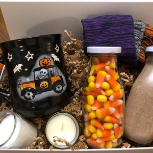 Puede incluir: Una caja de regalo blanca llena de art&iacute;culos con tem&aacute;tica de Halloween, que incluyen una taza negra con un esqueleto conduciendo un autom&oacute;vil, un frasco de vidrio lleno de ma&iacute;z dulce, una botella de vidrio llena de polvo marr&oacute;n, dos velas blancas peque&ntilde;as y un par de calcetines morados y grises.