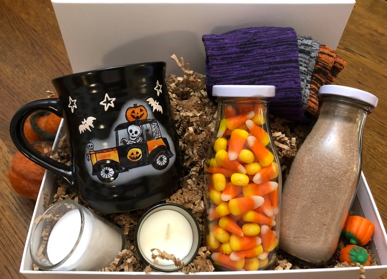 Puede incluir: Una caja de regalo blanca llena de art&iacute;culos con tem&aacute;tica de Halloween, que incluyen una taza negra con un esqueleto conduciendo un autom&oacute;vil, un frasco de vidrio lleno de ma&iacute;z dulce, una botella de vidrio llena de polvo marr&oacute;n, dos velas blancas peque&ntilde;as y un par de calcetines morados y grises.