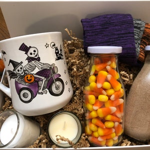 Puede incluir: Una caja de regalo con tem&aacute;tica de Halloween llena de una taza de cer&aacute;mica blanca con un esqueleto montando una motocicleta, dos velas, un frasco de vidrio lleno de ma&iacute;z dulce y una botella de vidrio llena de una sustancia marr&oacute;n.