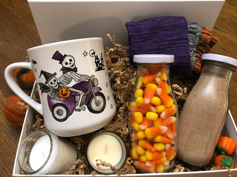 Puede incluir: Una caja de regalo con tem&aacute;tica de Halloween llena de una taza de cer&aacute;mica blanca con un esqueleto montando una motocicleta, dos velas, un frasco de vidrio lleno de ma&iacute;z dulce y una botella de vidrio llena de una sustancia marr&oacute;n.