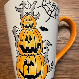 Puede incluir: Una taza de cer&aacute;mica blanca con asa naranja presenta un dise&ntilde;o de Halloween. Tres calabazas talladas apiladas con caras naranjas y negras est&aacute;n rodeadas de fantasmas y una telara&ntilde;a.