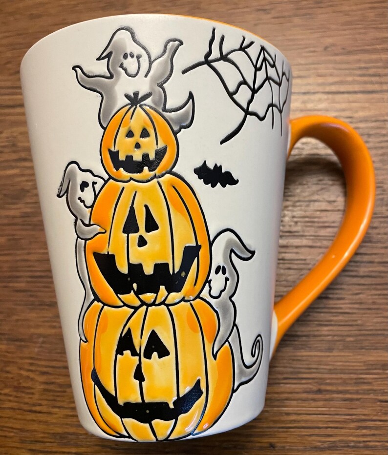 Puede incluir: Una taza de cer&aacute;mica blanca con asa naranja presenta un dise&ntilde;o de Halloween. Tres calabazas talladas apiladas con caras naranjas y negras est&aacute;n rodeadas de fantasmas y una telara&ntilde;a.